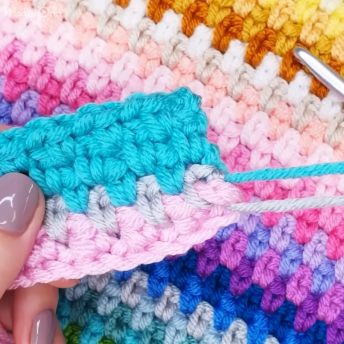 A hand holds a teal crochet row, transitioning to a pink and purple patterned fabric.
