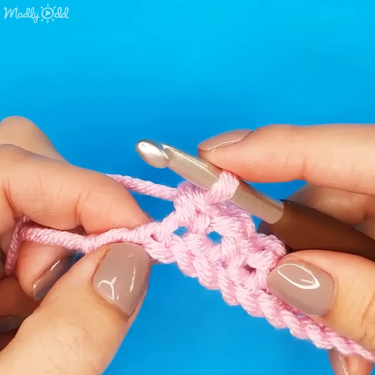 Hands with painted nails hold a crochet hook and pink yarn, demonstrating a crochet technique against a blue background.