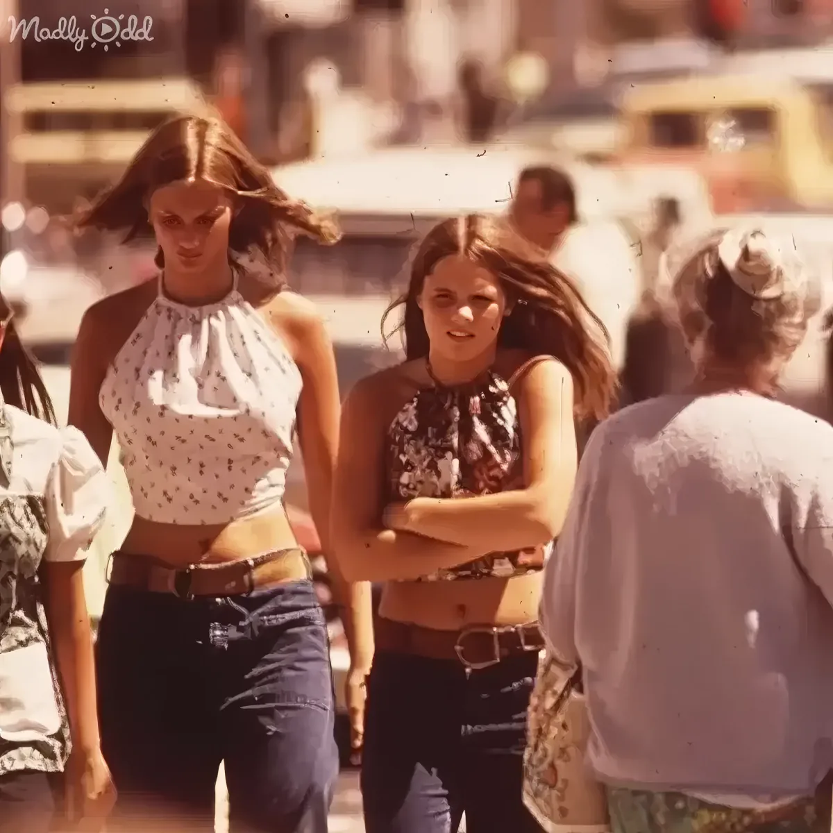 Two women in halter tops and jeans walk together, one with long hair, the other with arms crossed.