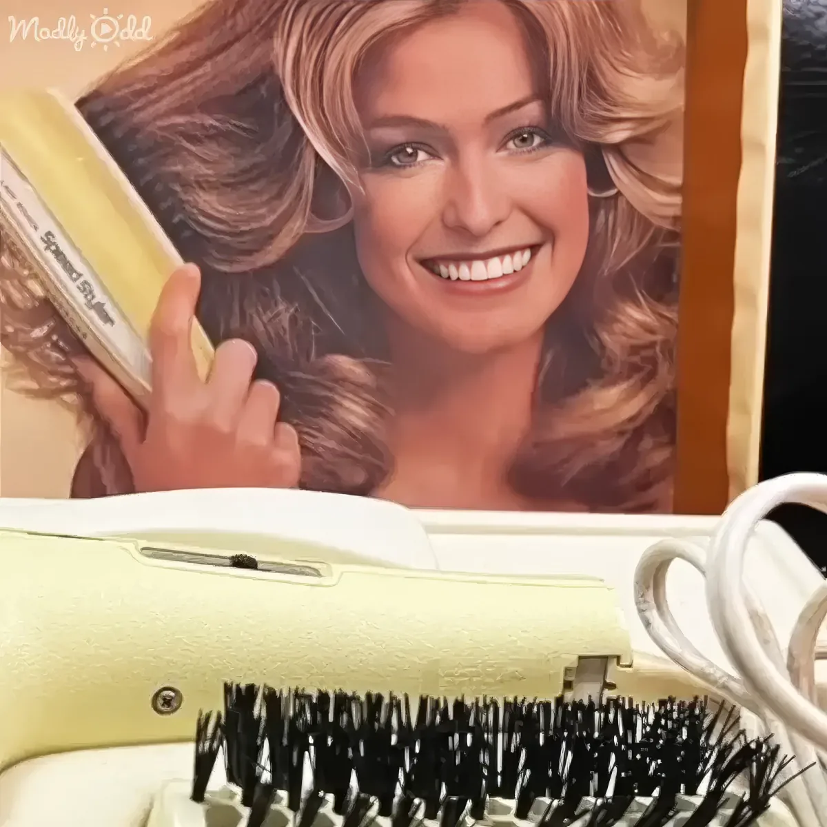 A vintage Speed Styler hair dryer with a brush attachment sits in front of a poster of a smiling woman holding a similar...