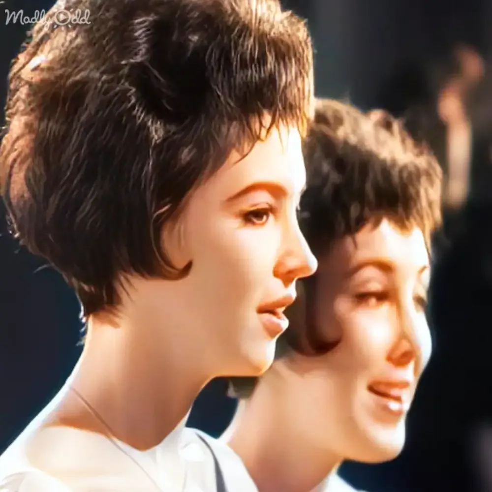 Two women with short, dark hair, one in white, both looking to the side, in a dimly lit setting.