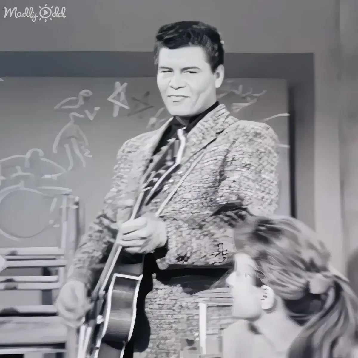A man in a patterned suit holds a guitar, smiling, with a chalkboard behind him.
