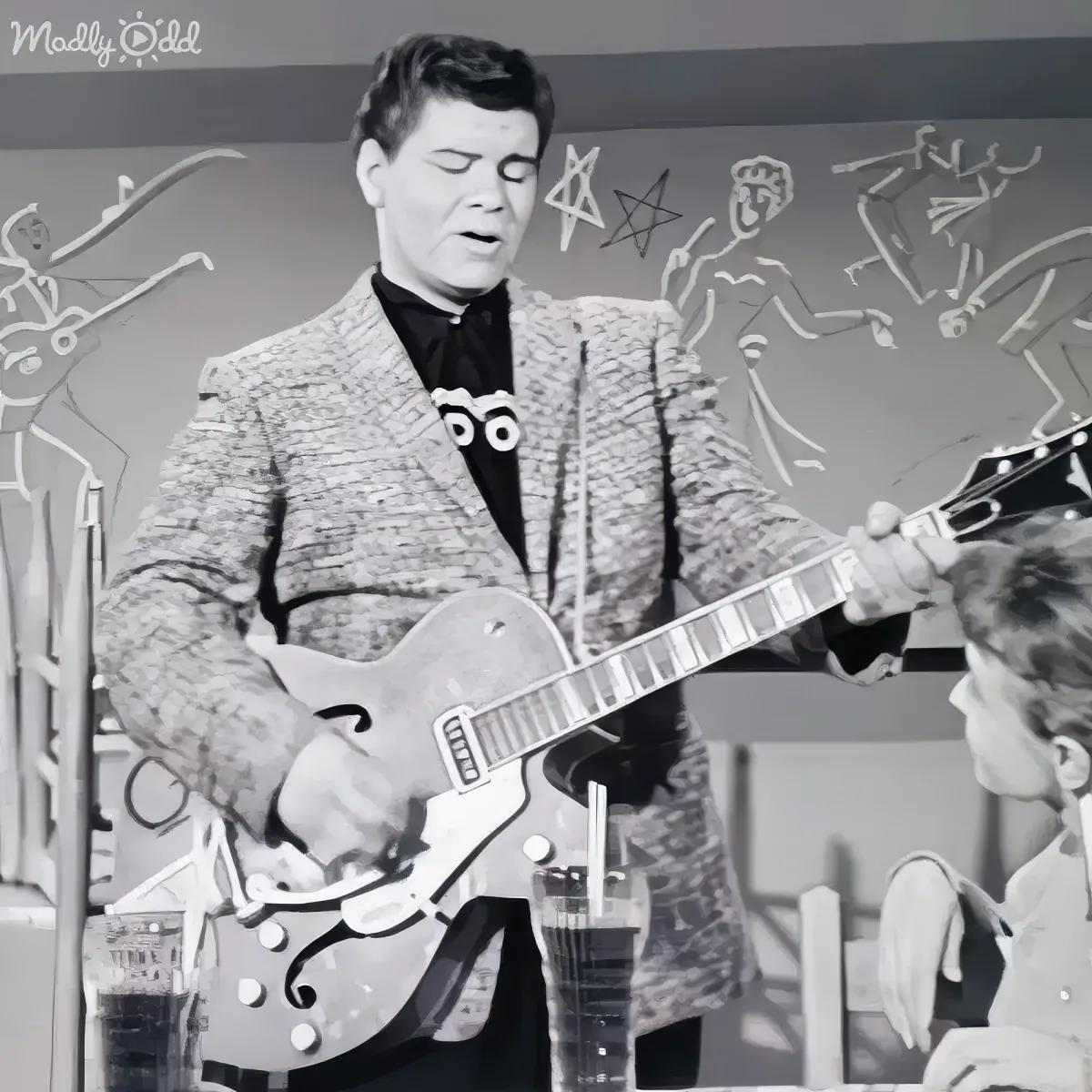 A man in a textured jacket plays a guitar on stage, with a microphone and a seated person nearby.