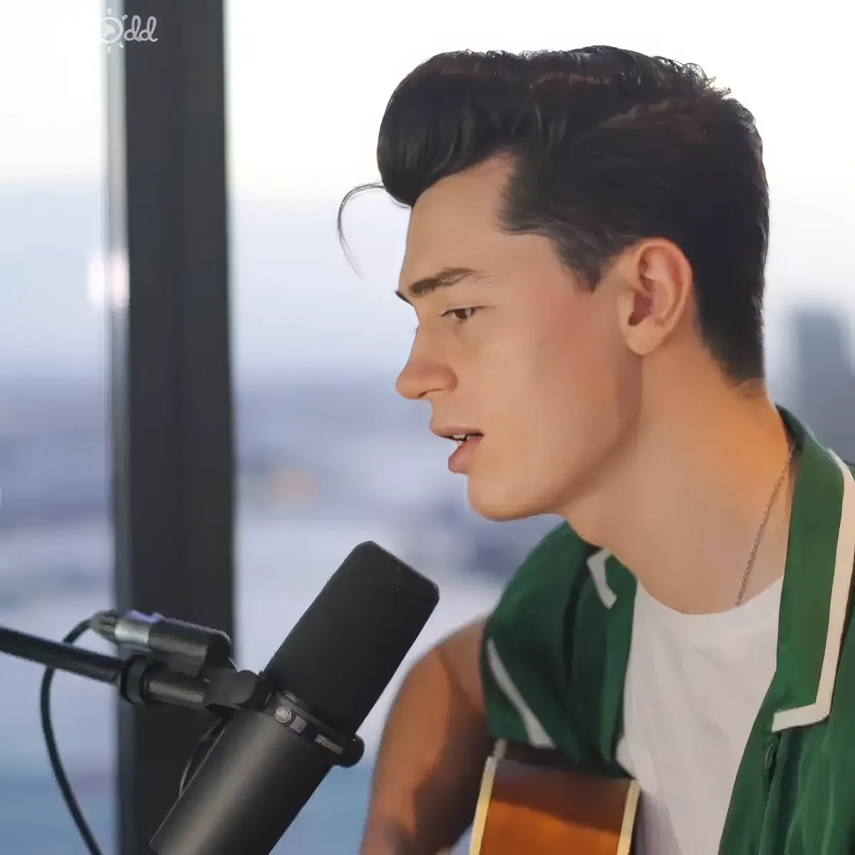 A man in a green jacket sings into a microphone, with a cityscape in the background.