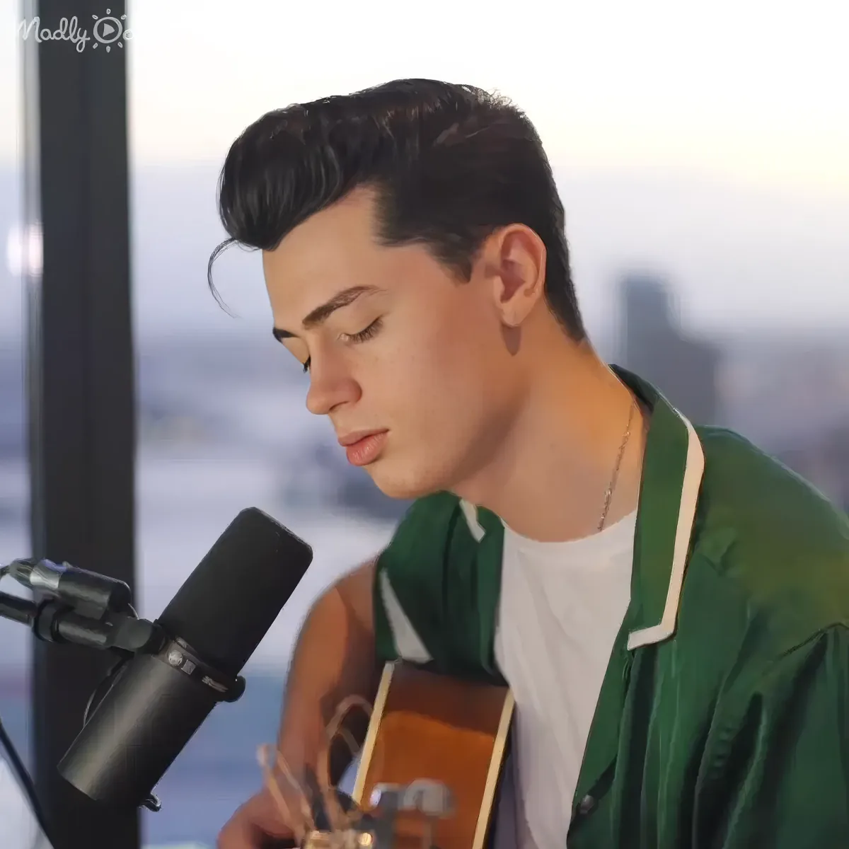 A person in a green jacket plays an acoustic guitar near a microphone, with a cityscape in the background.