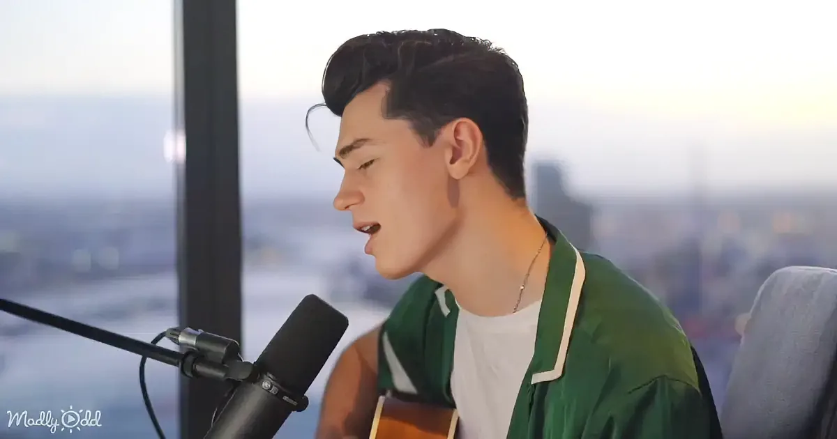 A person in a green jacket sings into a microphone, holding a guitar, with a cityscape in the background.