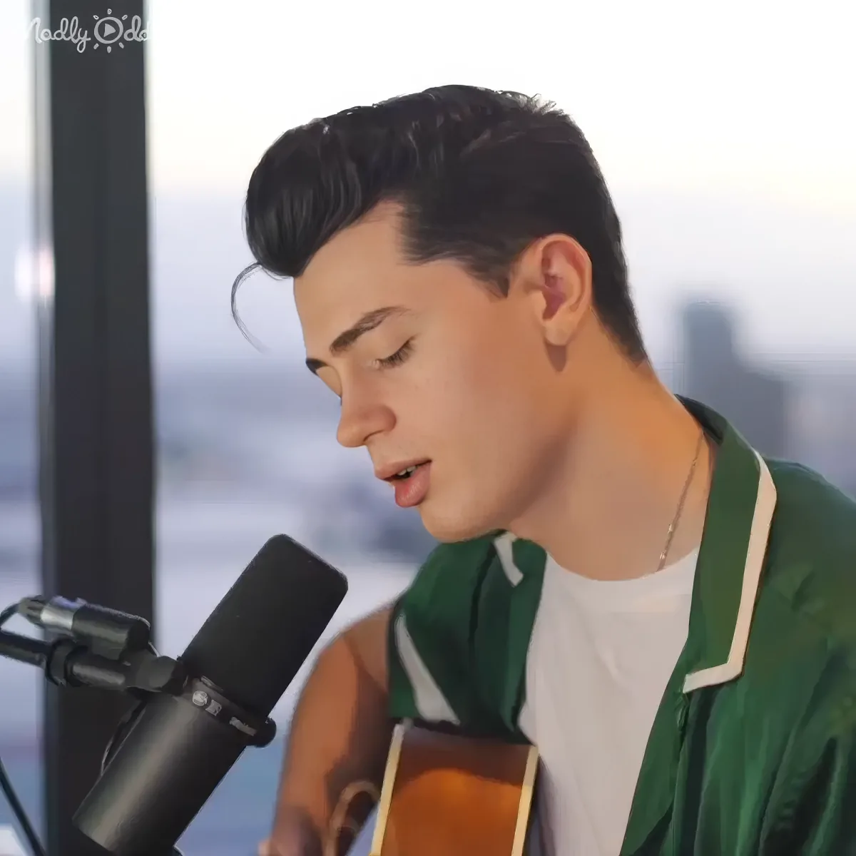 A person in a green jacket sings into a microphone while playing an acoustic guitar.