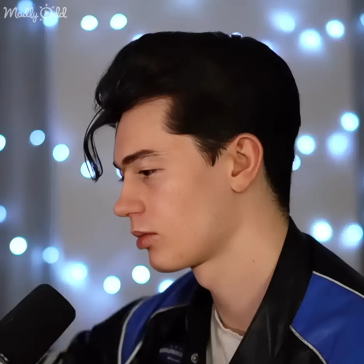 A person with dark hair and a black jacket with blue accents, looking down with a microphone in front of them.