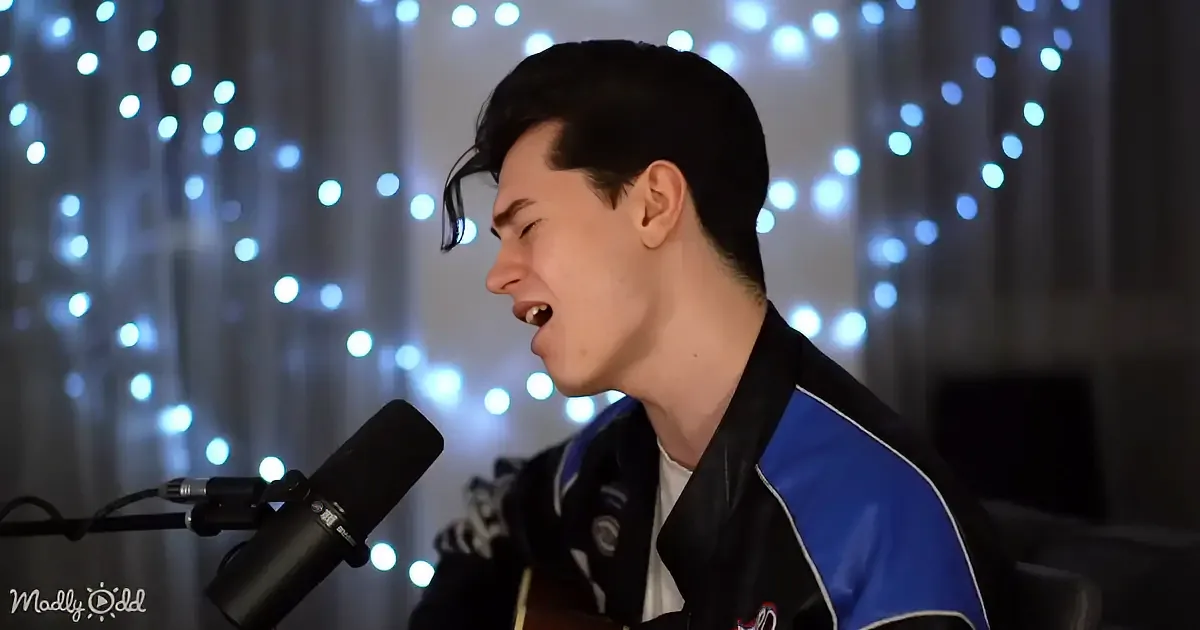 A person singing into a microphone, wearing a black and blue jacket, with string lights in the background.