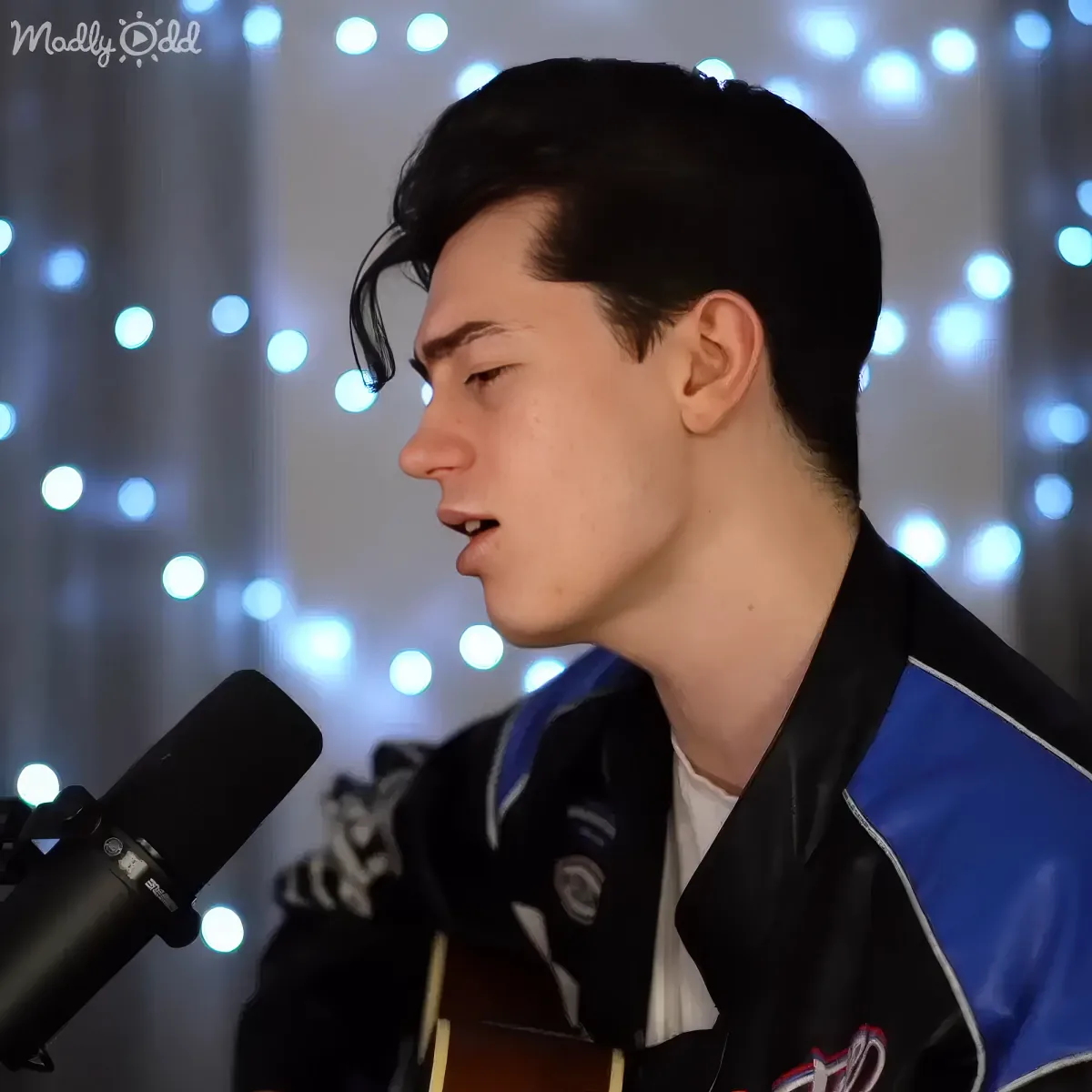 A person singing into a microphone, wearing a black and blue jacket, with string lights in the background.