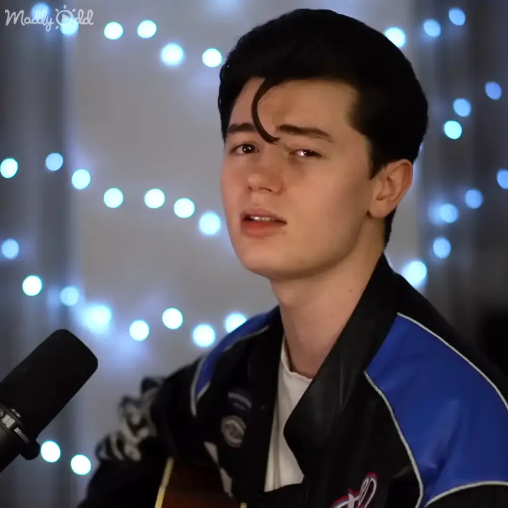 A young man with dark hair, wearing a black and blue jacket, sings into a microphone in front of a string of lights.