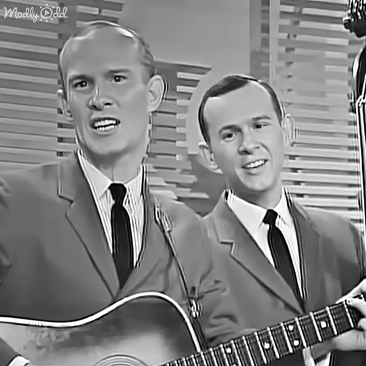 Two men in suits play guitars and sing, one holding a microphone, in a vintage black-and-white setting.