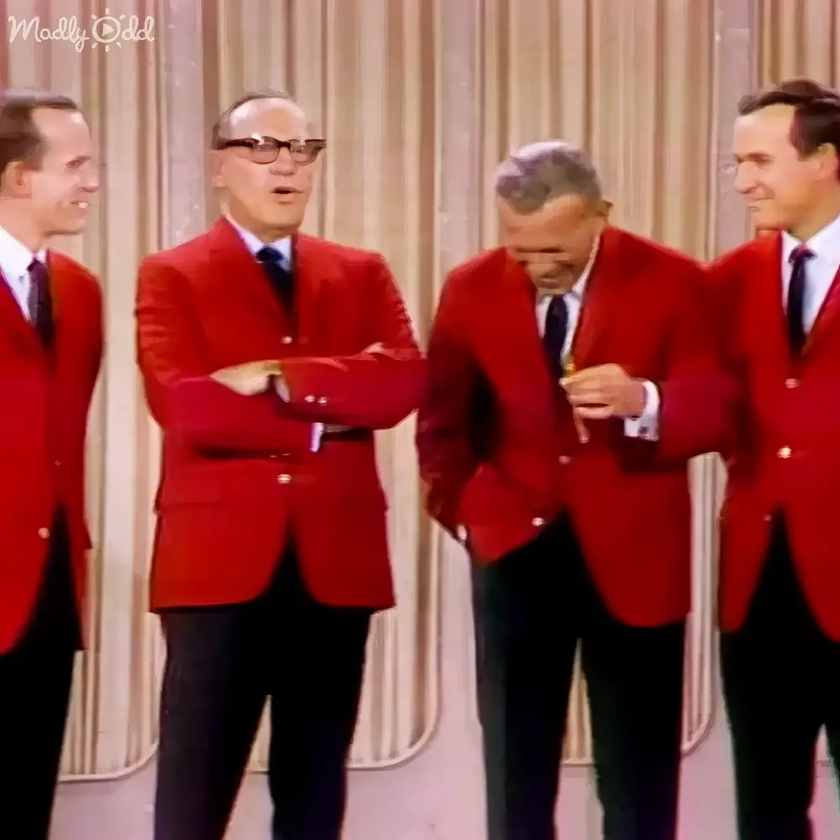 Four men in red jackets and black pants stand together, one adjusting his jacket.