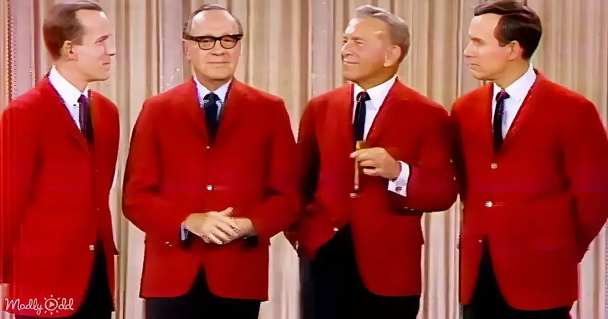 Four men in red velvet jackets stand together, smiling and holding drinks, against a curtain backdrop.