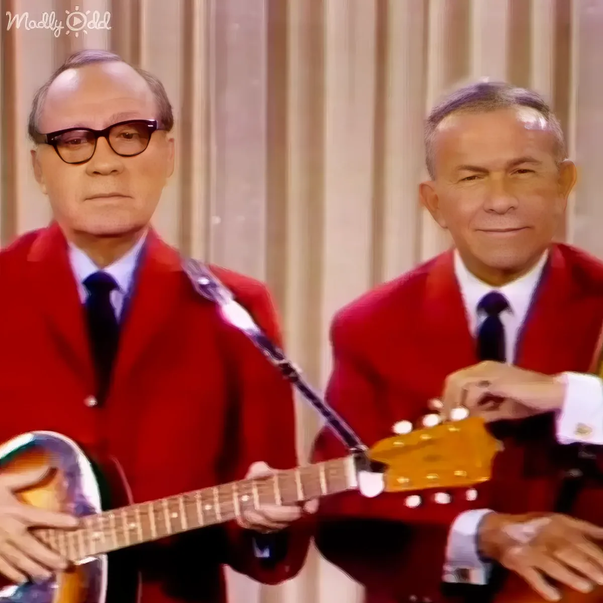 Two men in red suits play banjos, one wearing glasses, against a beige curtain backdrop.