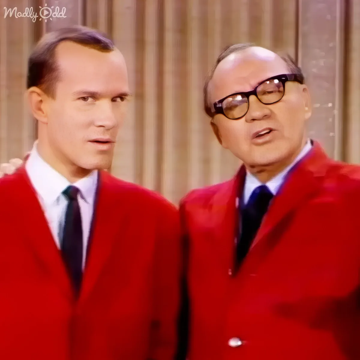 Two men in red suits and ties stand side by side, one wearing glasses, against a curtain backdrop.