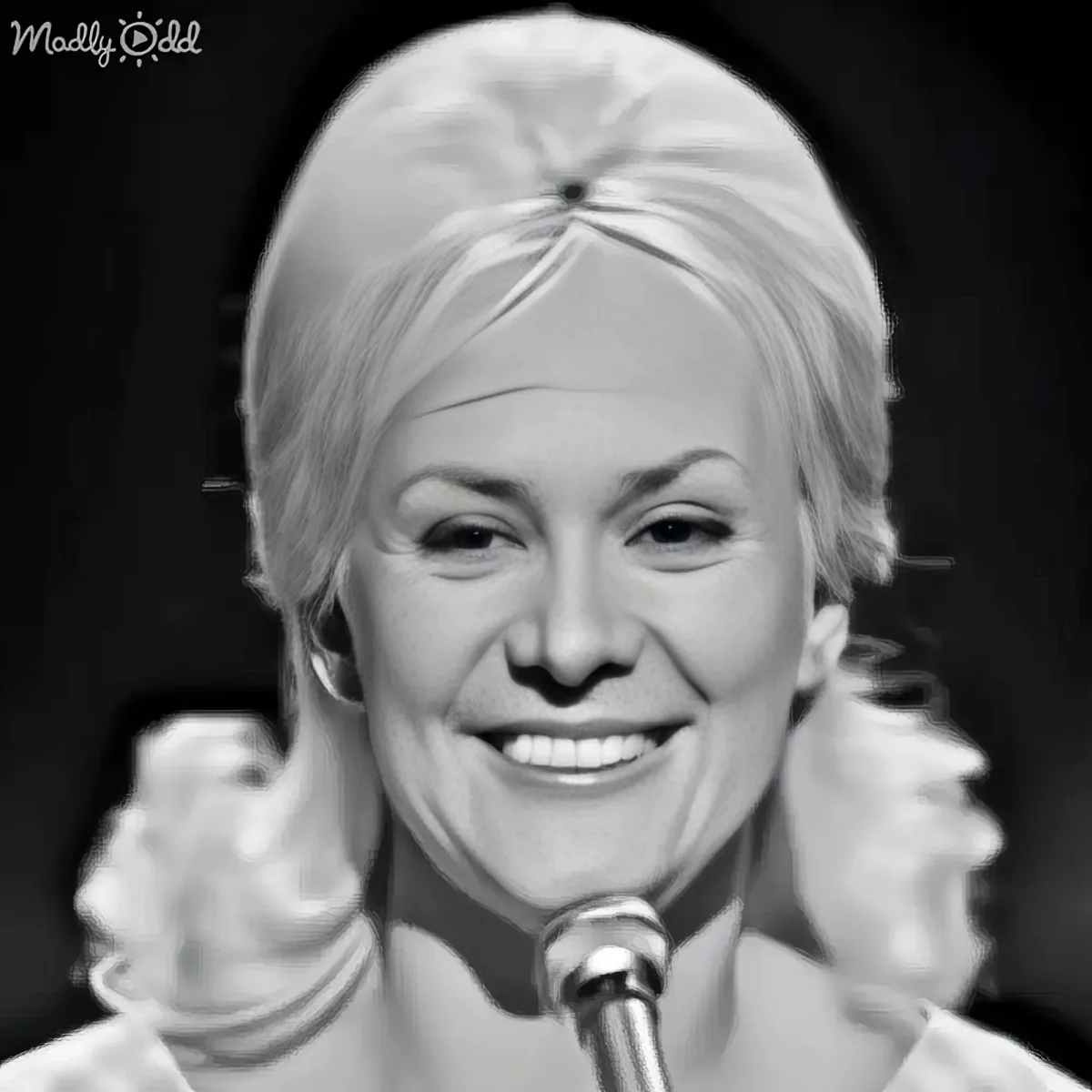 A smiling woman with blonde hair tied back, wearing a white top, holding a microphone.
