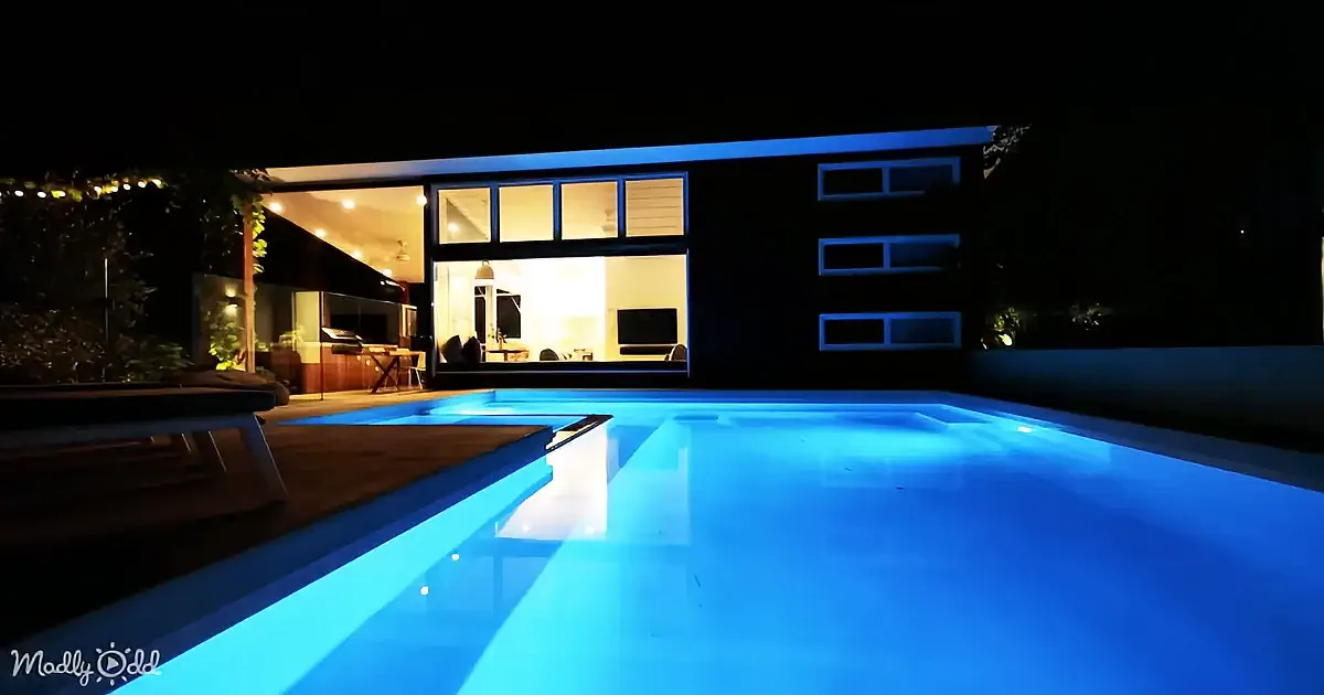 A glowing blue pool reflects the illuminated interior of a modern house at night.