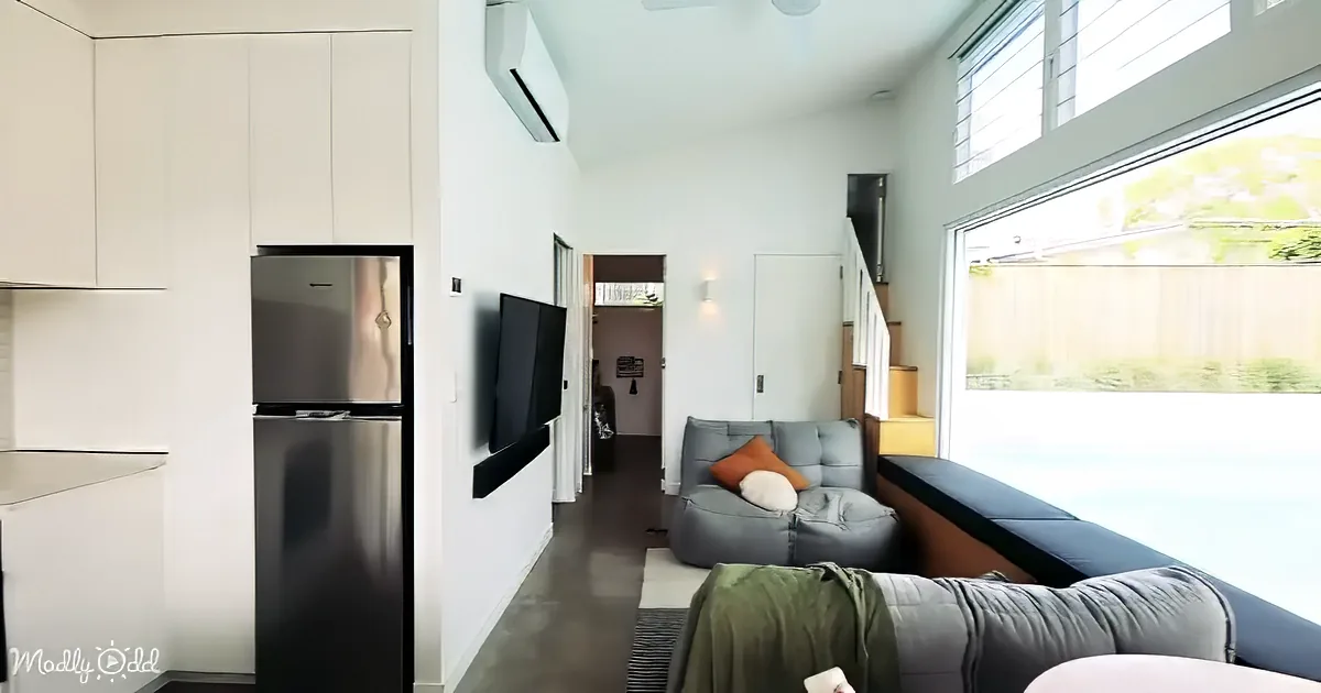 A modern living space with a gray sofa, wall-mounted TV, and stainless steel refrigerator in a bright, open-plan area.
