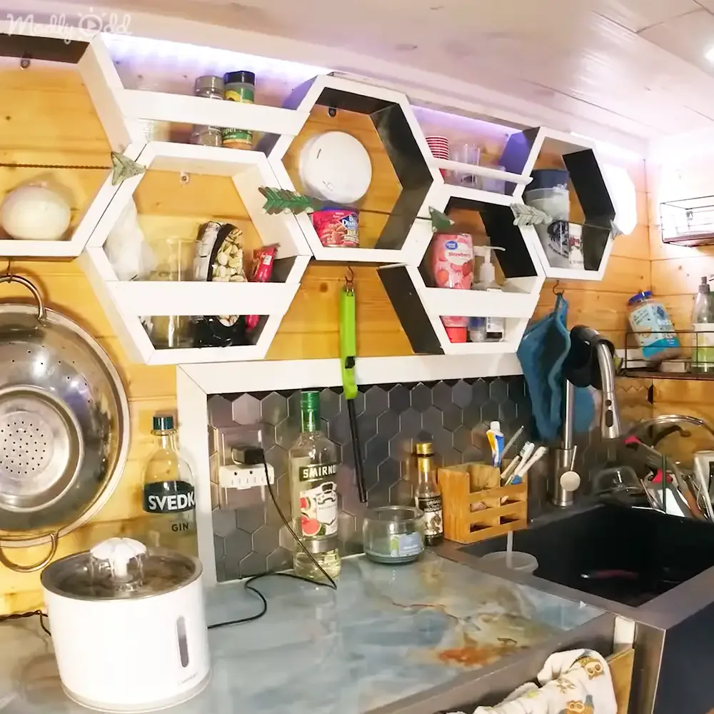 A kitchen with hexagonal shelves, wooden walls, and various items like spices, utensils, and bottles.