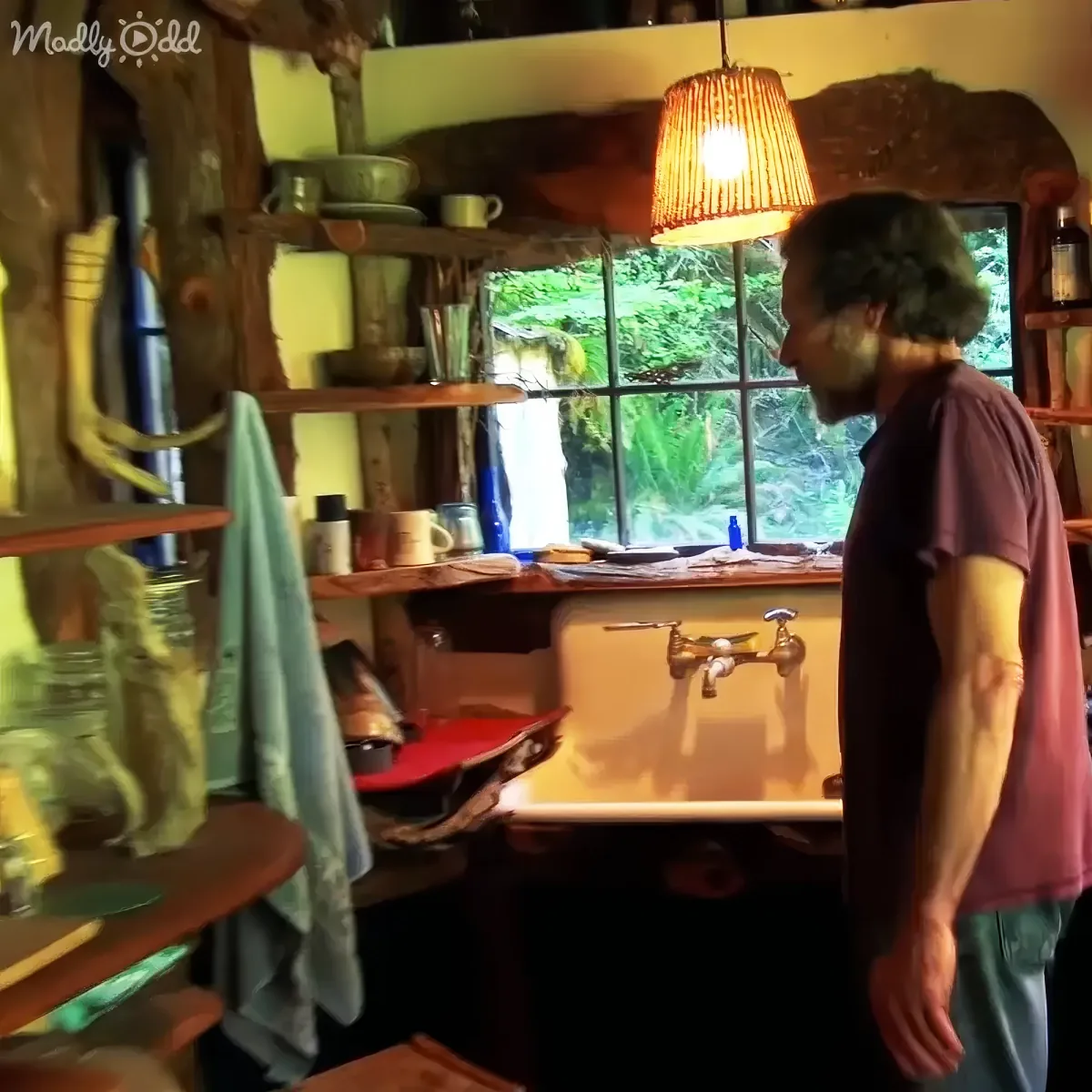 A man in a red shirt stands in a rustic kitchen with shelves, a window, and a hanging light.