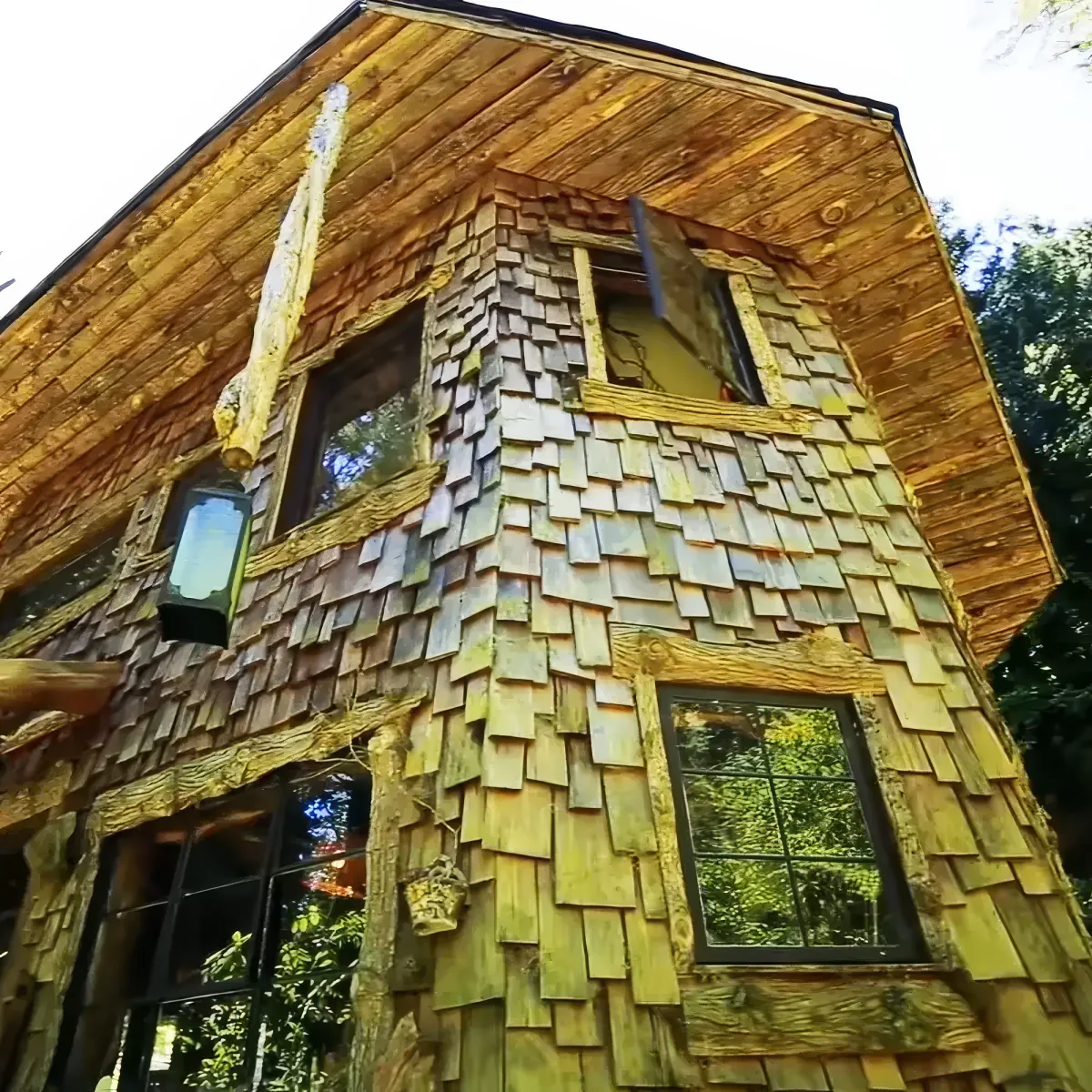 A rustic wooden house with shingles and large windows, surrounded by greenery.