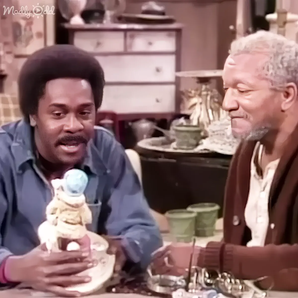 A man in a blue shirt holds a figurine while sitting at a table with another man in a white shirt.