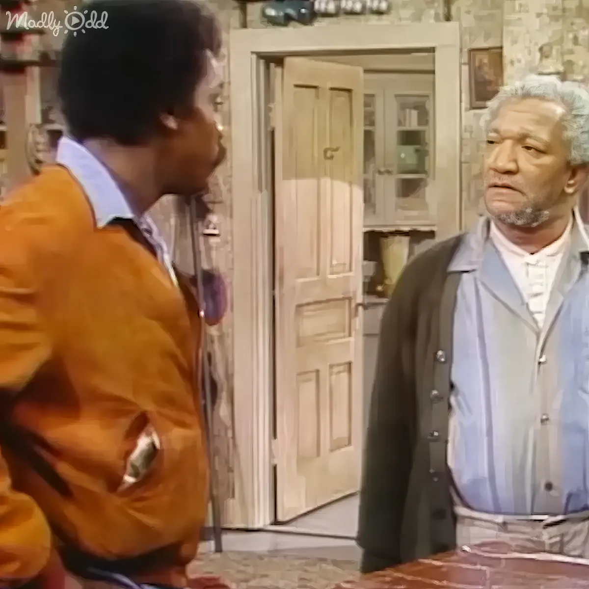 A man in an orange jacket stands facing another man in a kitchen, both appearing to be in conversation.