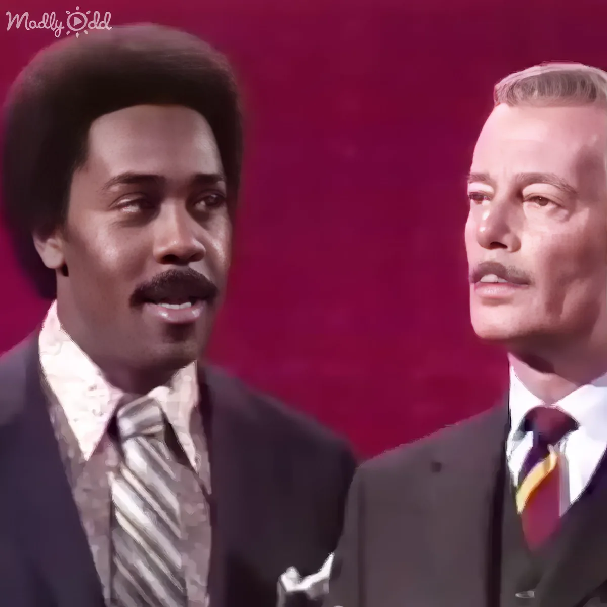 Two men in suits, one with a mustache, standing against a red background.