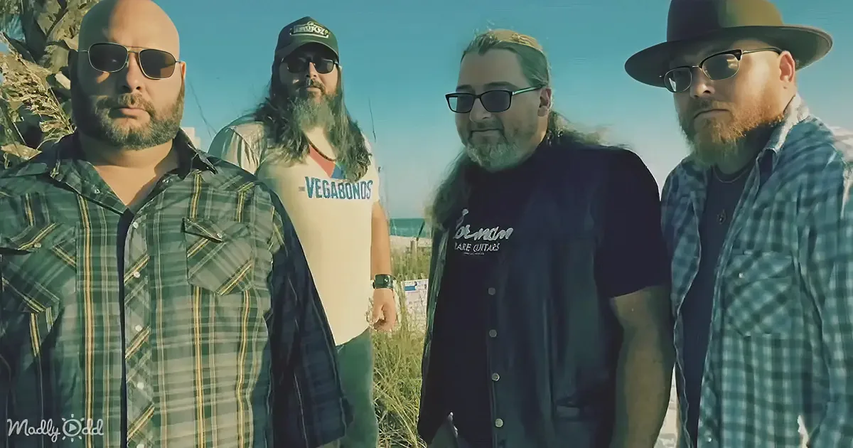 Four men in casual attire stand outdoors, one wearing a "Vagabonds" shirt, under a clear sky.