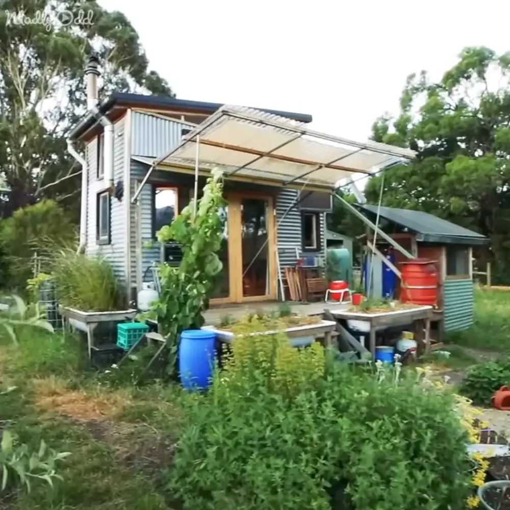 A small, eco-friendly house with a garden, solar panels, and various containers for plants and storage.