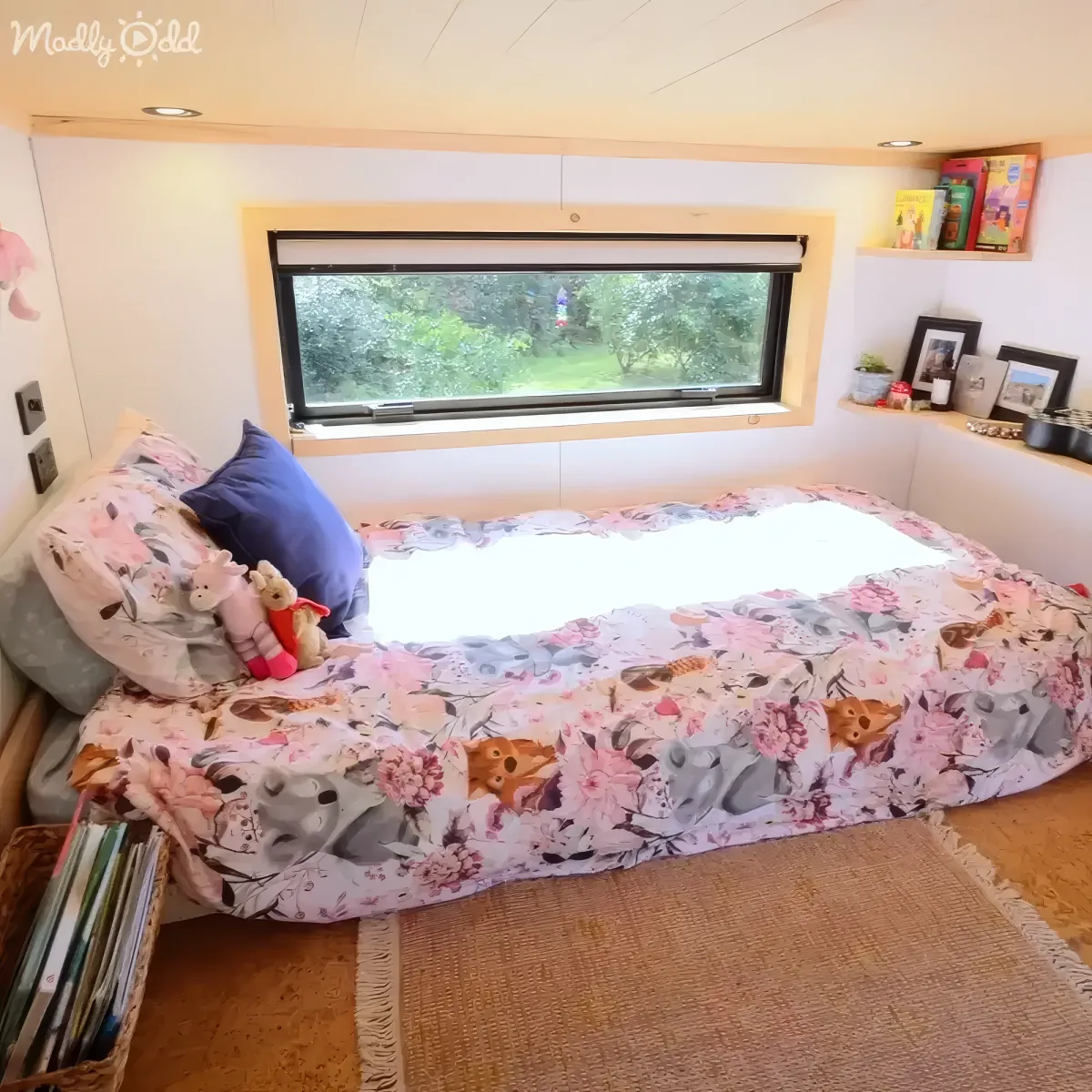A cozy bedroom with a floral bedspread, stuffed animals, and a window showing greenery outside.