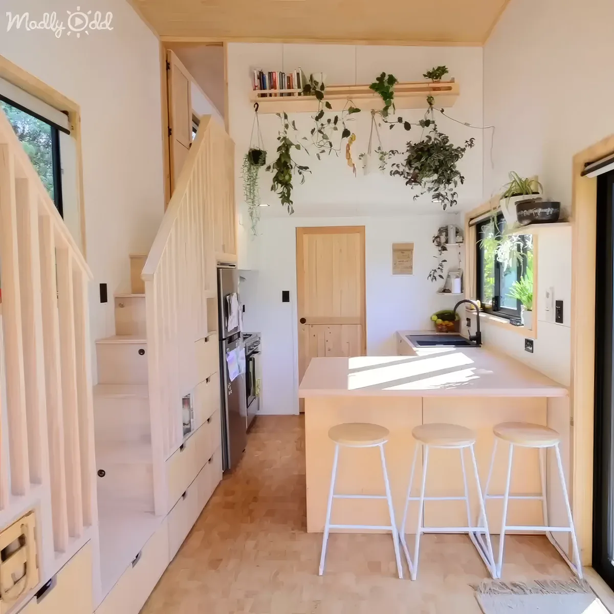 A cozy kitchen with a central island, stools, hanging plants, and a wooden staircase leading to a loft.