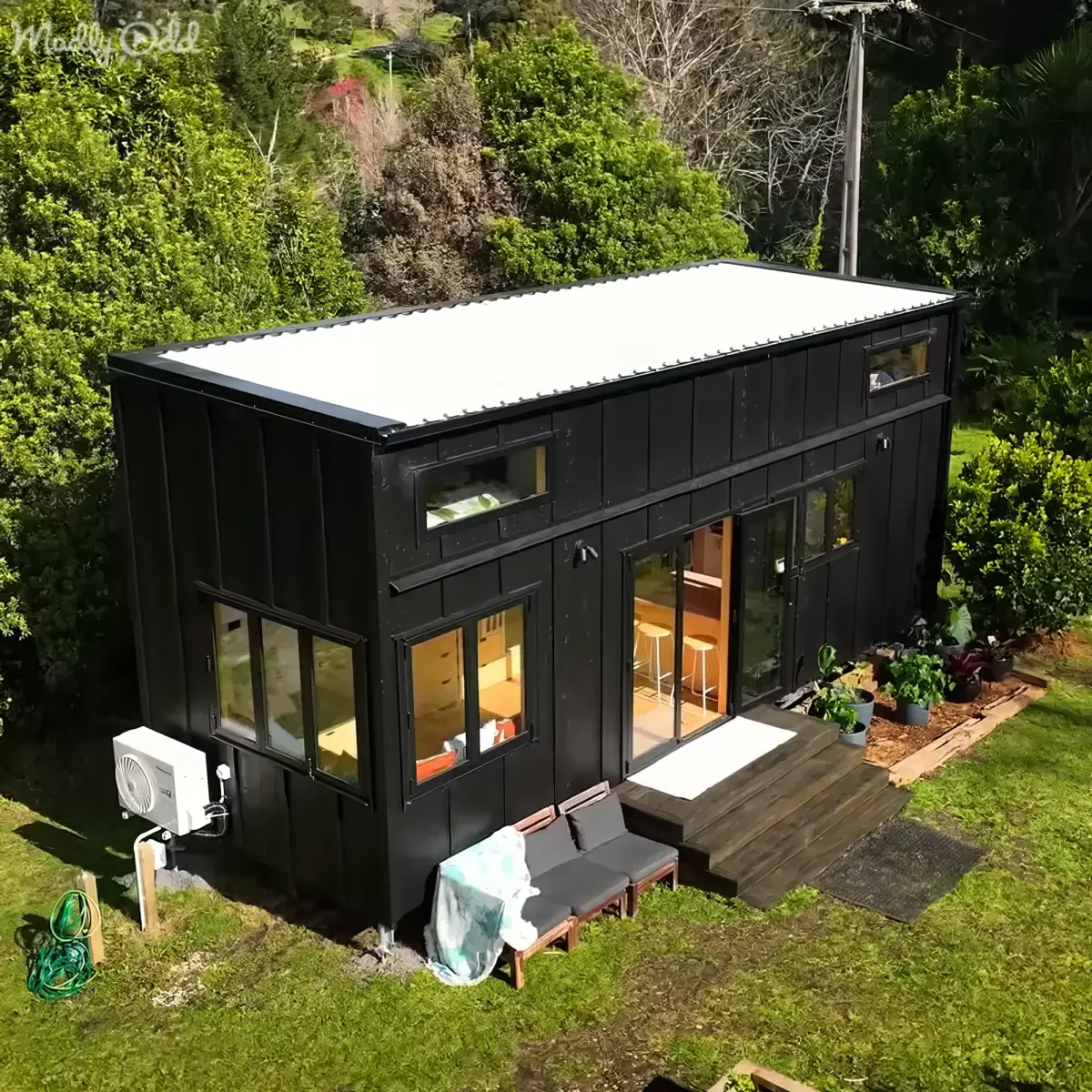 A modern black tiny house with a white roof sits surrounded by lush greenery, featuring large windows and a wooden deck.