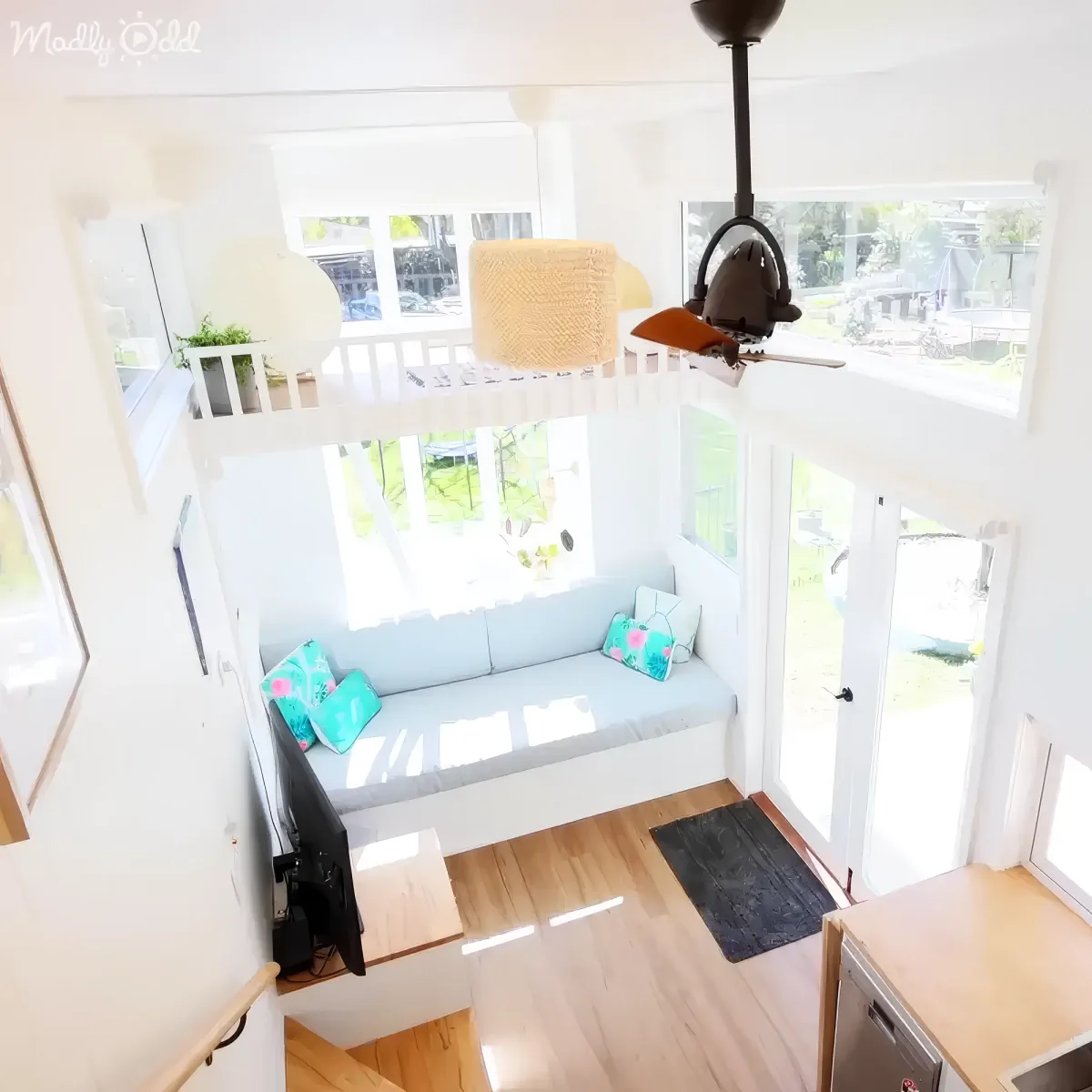 A cozy, bright living space with a light blue couch, colorful pillows, a ceiling fan, and large windows.