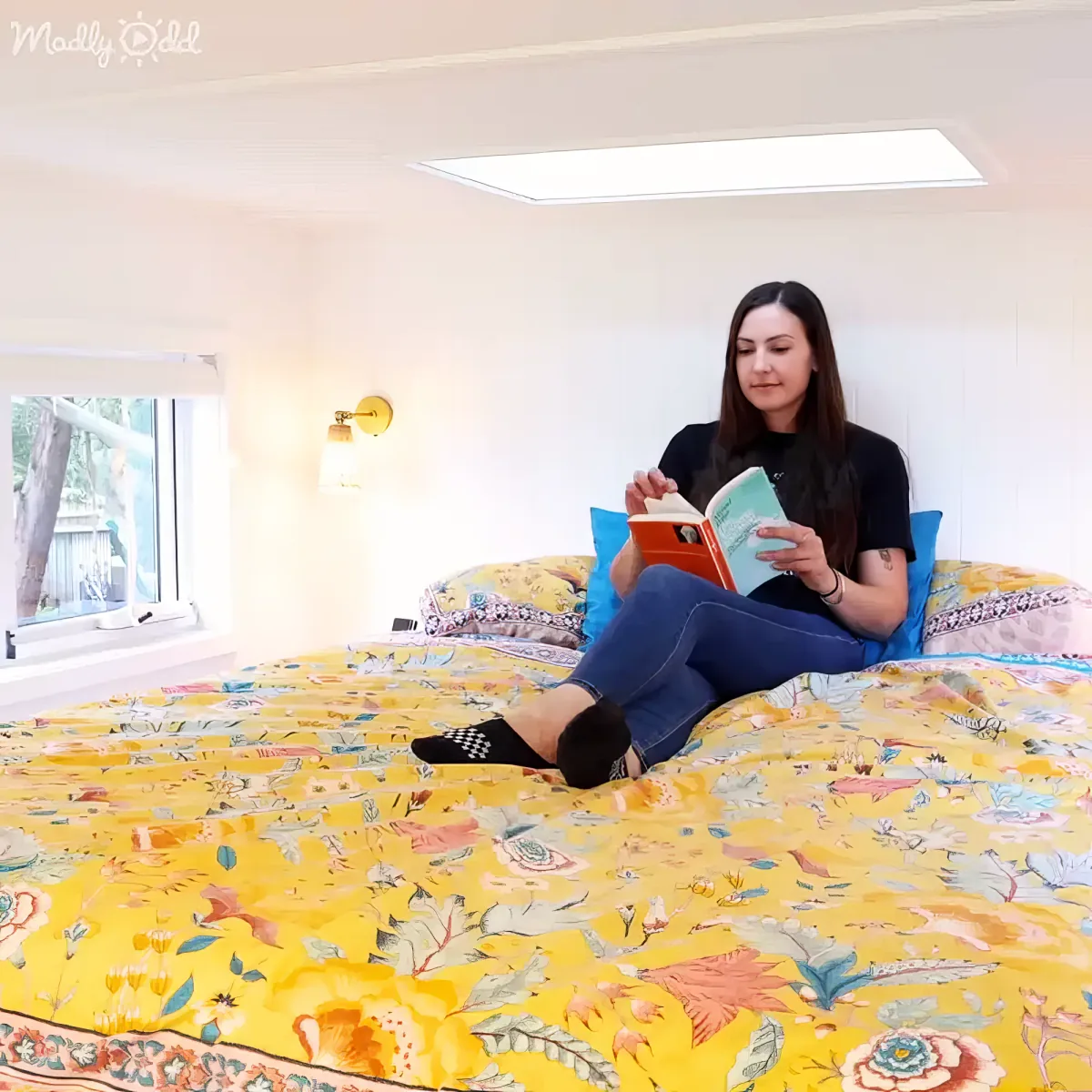 A woman sits on a bed with a yellow floral blanket, reading a book.