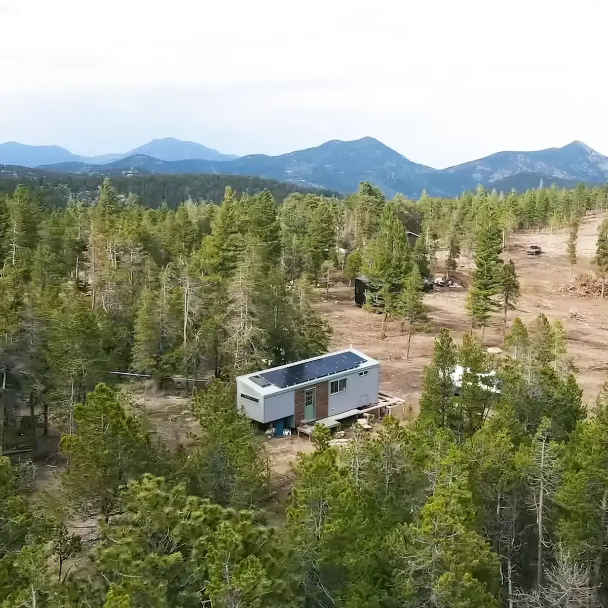 A small house with solar panels sits surrounded by dense trees, with mountains in the background.