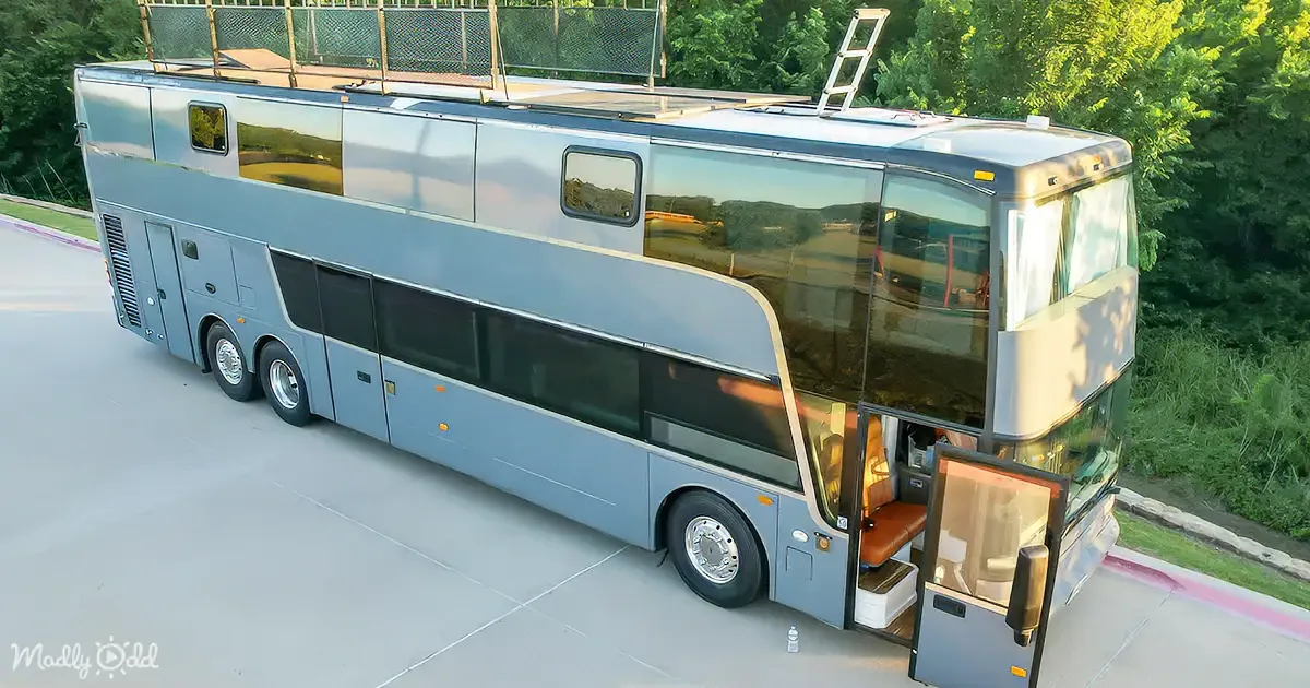A large, modern, two-story bus with a ladder on top is parked on a road near a fence and trees.