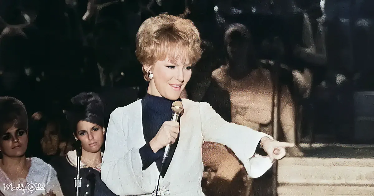A woman in a white outfit holds a microphone, pointing to the right, with a crowd in the background.