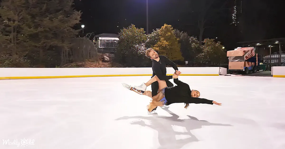 A couple performs a synchronized ice skating move on a rink at night.