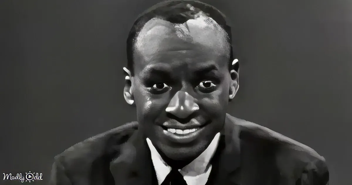 A man in a suit and tie smiles, looking directly at the camera against a plain background.