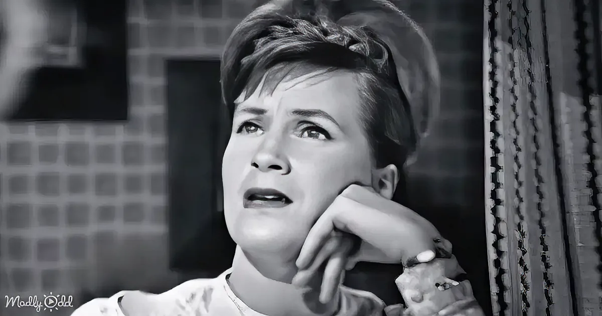 A woman with short hair, wearing a white blouse, rests her head on her hand, looking upwards with a concerned expression.