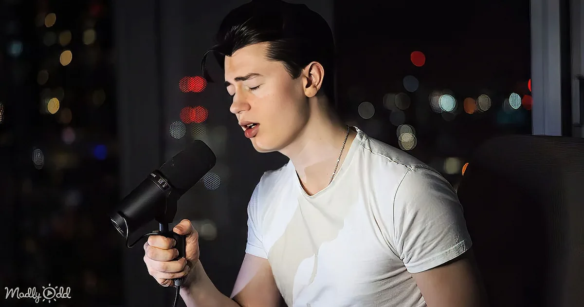 A person in a white shirt holds a microphone, singing with a cityscape in the background.