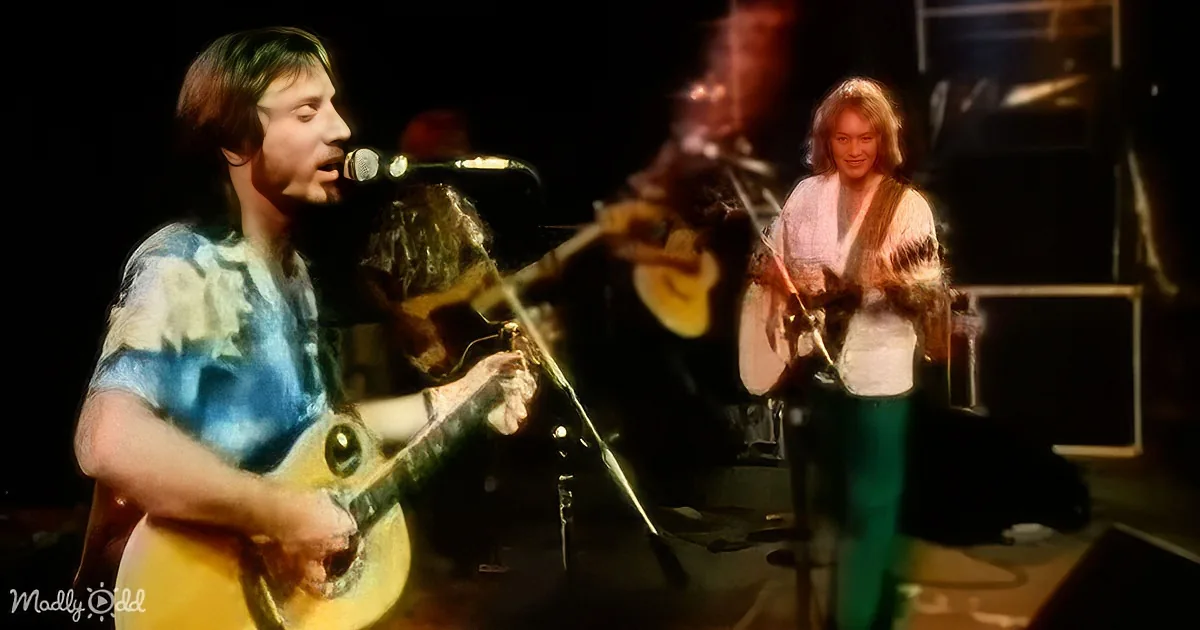 A man in a tie-dye shirt plays guitar on stage, with another musician in the background.