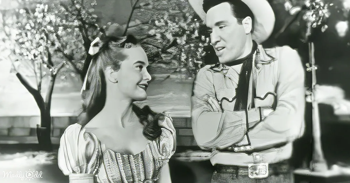 A woman in a vintage dress smiles at a man in a cowboy outfit, both seated outdoors.