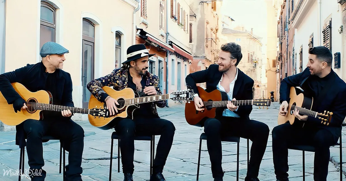 Four men in suits play guitars on a cobblestone street in a sunlit European town.