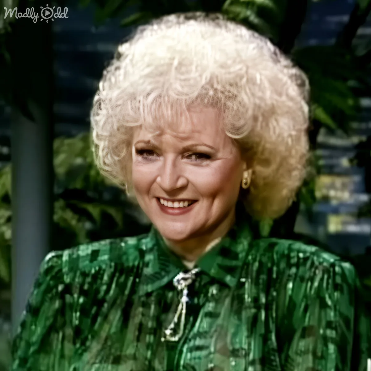 A smiling woman with curly blonde hair, wearing a green patterned top and a necklace, stands in front of a blurred...