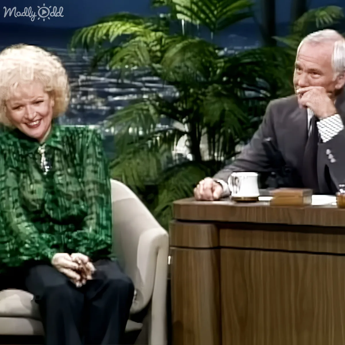 A woman in a green patterned top sits on a chair, smiling, while a man in a suit leans on a desk, holding his hand to his...