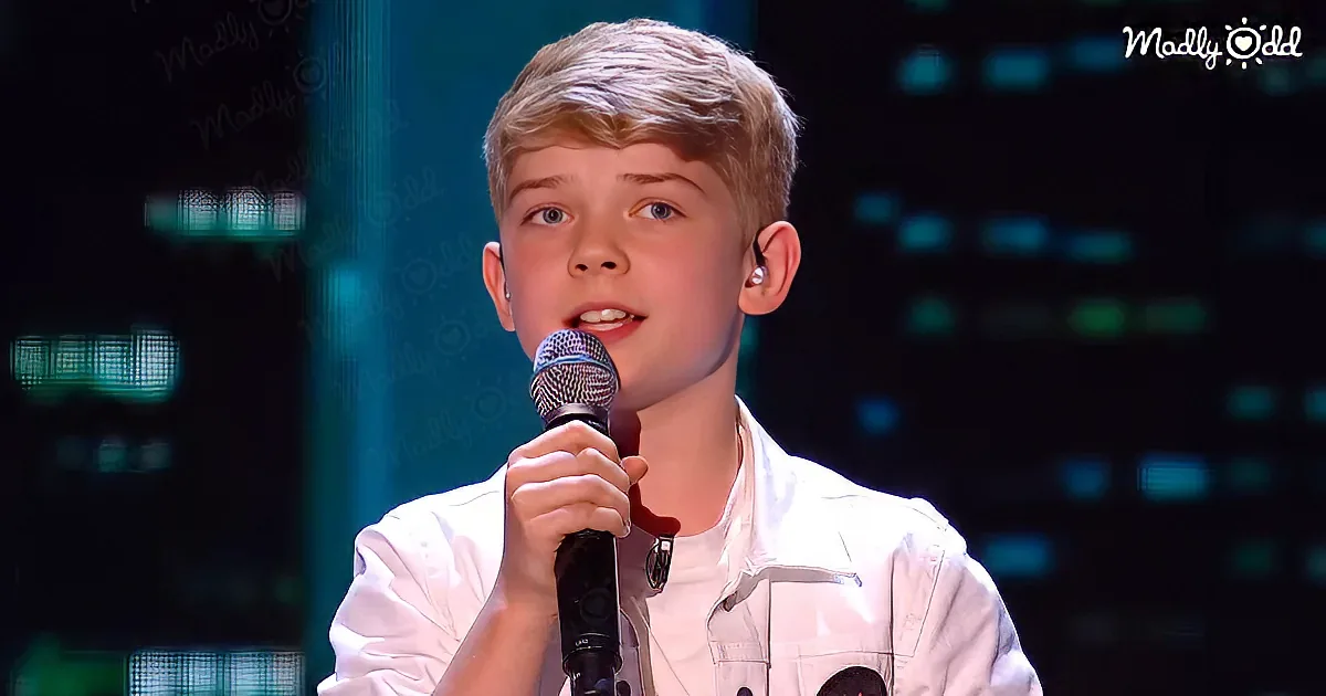 A young boy in a white shirt holds a microphone, singing on stage with a dark background.