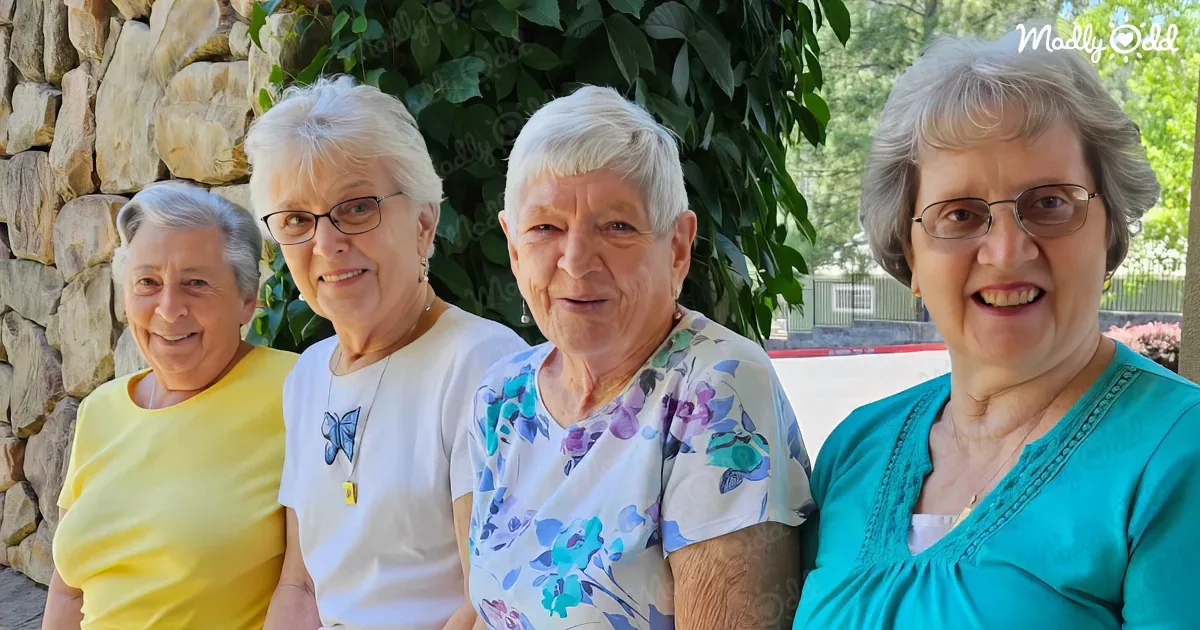 Four elderly women in colorful shirts smile and pose together outdoors near a stone wall and greenery.